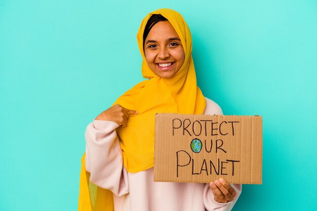 Jovem mulher muçulmana segurando um proteger nosso planeta isolado na parede azul pessoa apontando com a mão para um espaço de cópia de camisa, orgulhosa e confiante