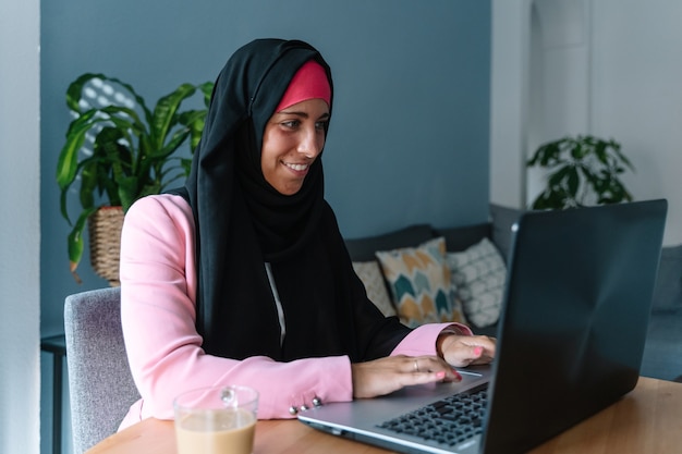 Jovem mulher muçulmana usando um hijab usando tecnologia dentro de casa. Vista lateral horizontal da mulher árabe, trabalhando com o laptop em casa. Tecnologia e estilo de vida das mulheres muçulmanas.