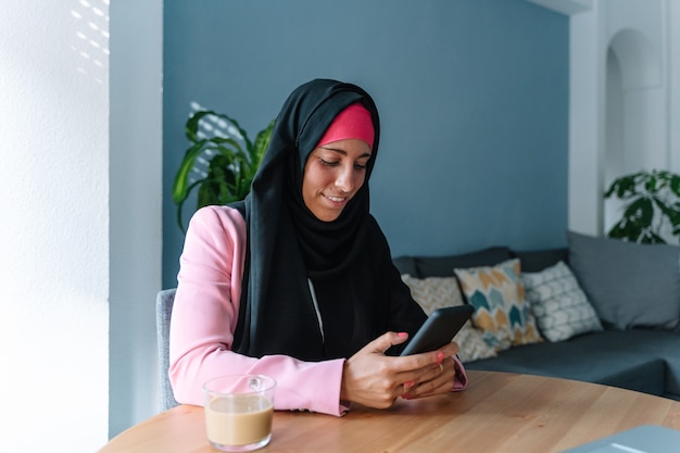 Jovem mulher muçulmana usando um hijab usando tecnologia dentro de casa. Vista panorâmica horizontal de mulher árabe trabalhando com smartphone em casa. Tecnologia e estilo de vida das mulheres muçulmanas.