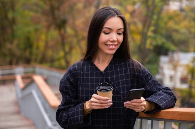 Jovem mulher muito sorridente de casaco com café para ir feliz usando o celular no parque