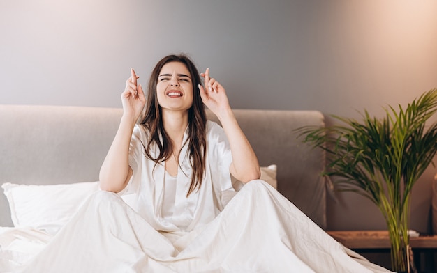 Jovem mulher na cama de pijama branco cruzou os dedos para dar sorte. de manhã ou à noite antes de um evento importante. a mulher está nervosa.