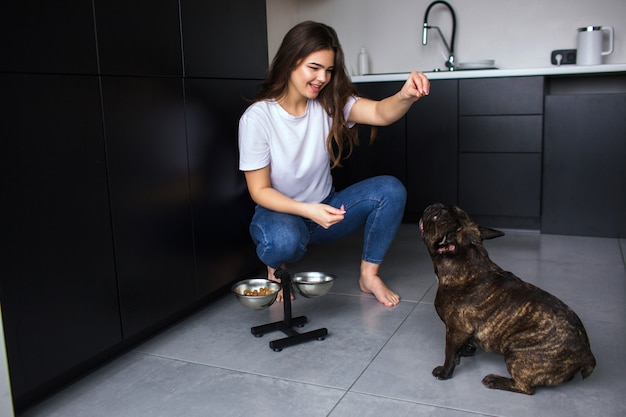 Jovem mulher na cozinha durante a quarentena. garota sentar e brincar com o bulldog francês. pet treinamento para fazer comandos e comer comida de cachorro depois disso.