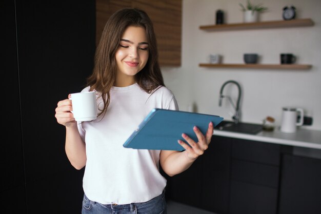 Jovem mulher na cozinha durante a quarentena. menina segura copo branco em uma mão e olha para tablet em outra. trabalhando em casa remota. conectados. tecnologias modernas.