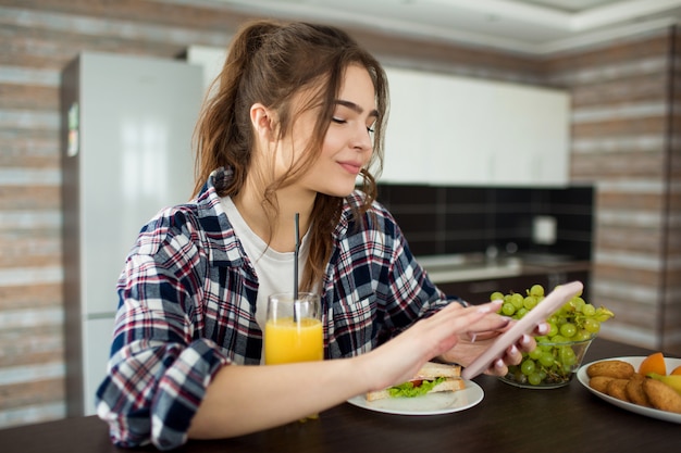 Foto jovem mulher na cozinha, tomando café da manhã ou almoço e usando o smartphone.