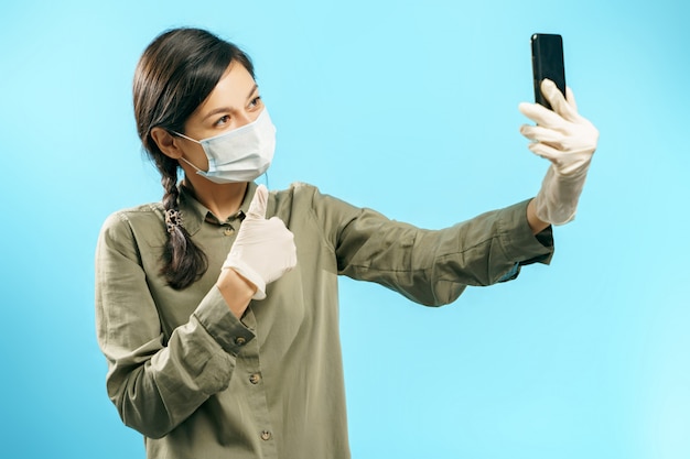 Jovem mulher na máscara médica protetora e luvas fazendo selfie ou vídeo chamada usando smartphone aparecendo polegar em azul.