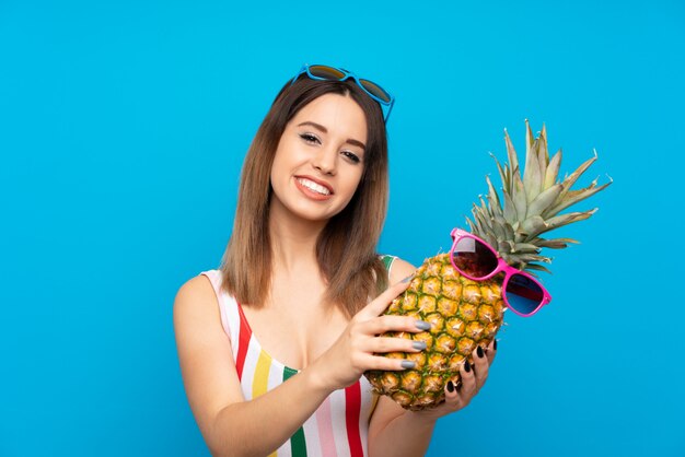 Jovem mulher nas férias de verão na parede azul, segurando um abacaxi com óculos de sol