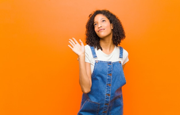 Jovem mulher negra bonita sorrindo alegremente e alegremente, acenando com a mão, dando as boas-vindas e cumprimentando-o ou dizendo adeus contra a parede laranja