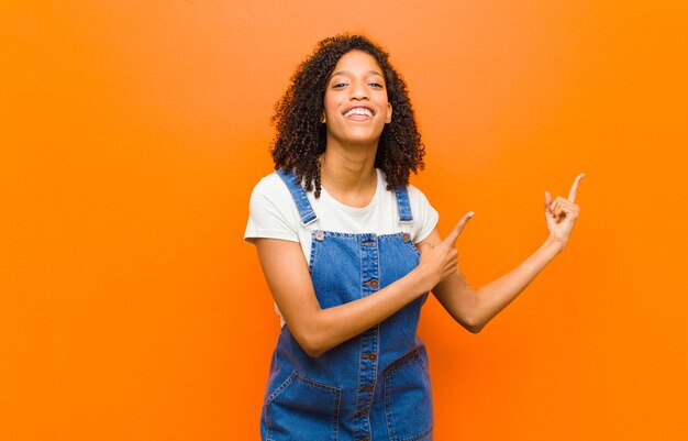 Jovem mulher negra bonita sorrindo alegremente e apontando para o lado e para cima com as duas mãos, mostrando o objeto no espaço da cópia contra a parede laranja