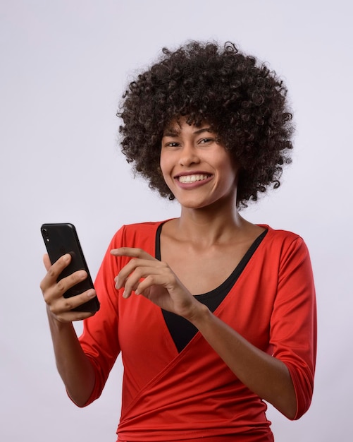 Foto jovem mulher negra brasileira segurando e olhando para um dispositivo de telefone celular sorrindo vestindo uma blusa vermelha