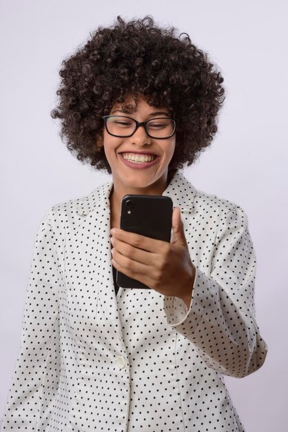 Foto jovem mulher negra brasileira segurando e olhando para um dispositivo de telefone celular sorrindo vestindo uma camisa branca