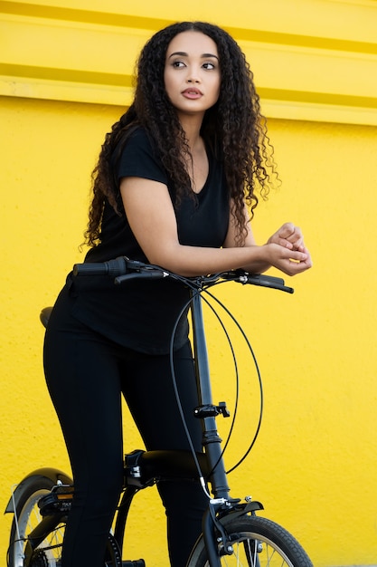 Jovem mulher negra com cabelo comprido e roupa preta em pé em sua bicicleta na frente de uma parede amarela