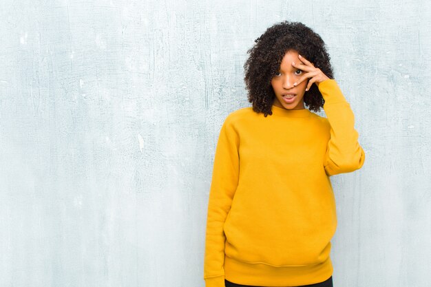 Jovem mulher negra olhando chocado, assustado ou aterrorizado, cobrindo o rosto com a mão e espreitando entre os dedos