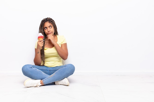 Jovem mulher negra pensando, sentindo-se duvidosa e confusa, com diferentes opções, imaginando qual decisão tomar. conceito de sorvete