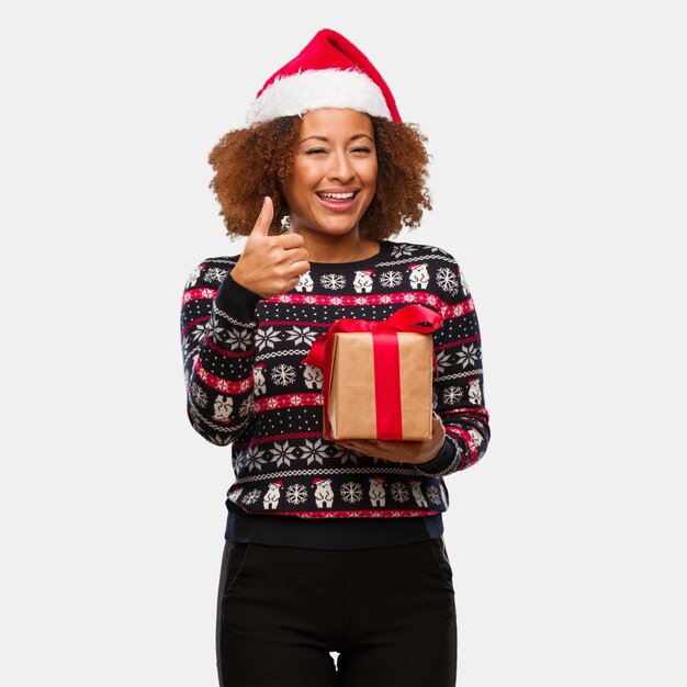 Jovem mulher negra segurando um presente no dia de natal, sorrindo e levantando o polegar para cima