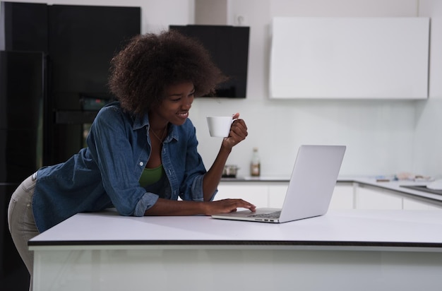 Jovem mulher negra sorridente usando computador e bebendo café no interior da cozinha moderna