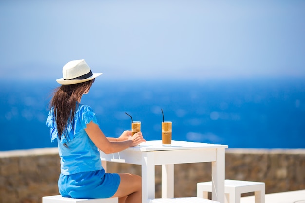 Jovem mulher no café ao ar livre, bebendo café frio, apreciando a vista para o mar.