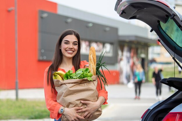 Jovem mulher no estacionamento carregando compras no porta-malas do carro Compras feitas com sucesso Mulher colocando malas no carro depois das compras