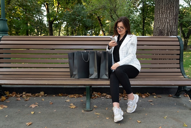 Jovem mulher no parque com sacolas de compras e telefone. Compras online. Descontos, promoções, black friday.