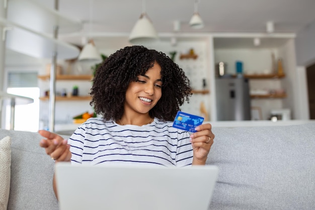 Jovem mulher no sofá fazendo compras online com cartão de débito Linda mulher africana usando computador portátil para compras online em casa