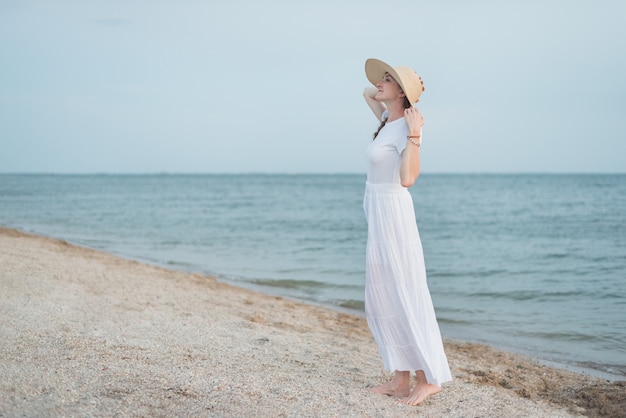 Jovem mulher no vestido branco longo e pé de chapéu na praia. Mar da noite.