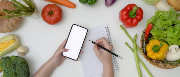 Jovem mulher observando o plano de dieta enquanto procurava informações no smartphone de tela em branco na mesa com legumes frescos