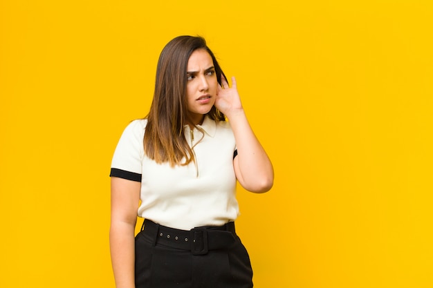 Jovem mulher olhando sério e curioso, ouvindo, tentando ouvir uma conversa secreta ou fofoca, bisbilhotando isolado na parede laranja