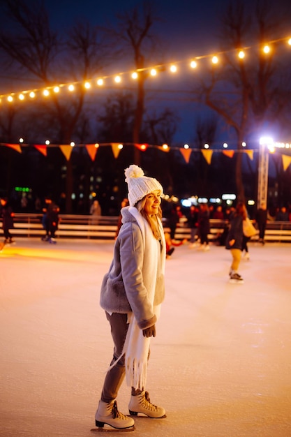 Jovem mulher patinando no gelo em um rinque em uma feira festiva de Natal à noite. Luzes ao redor.