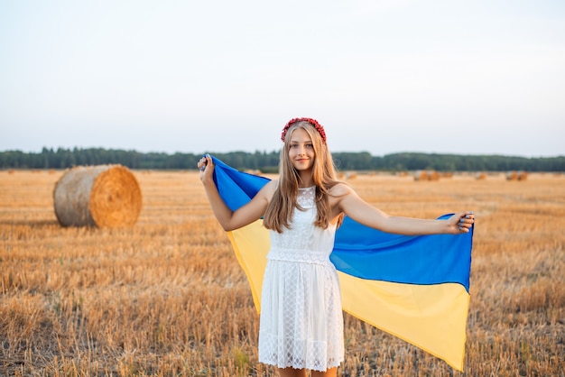Jovem mulher patriótica com uma bandeira da Ucrânia no campo de trigo colhido com alguns fardos de palha