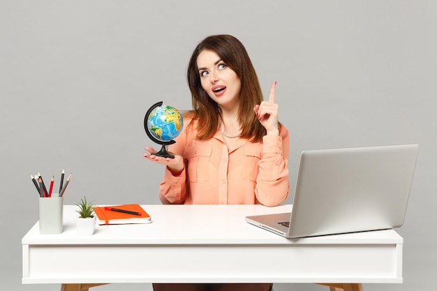 Jovem mulher pensativa, apontando o dedo indicador para cima, segurando o globo do mundo da terra sentar trabalho na mesa com o laptop pc isolado em fundo cinza. conceito de estilo de vida de carreira de negócios de realização. mock up espaço de cópia.