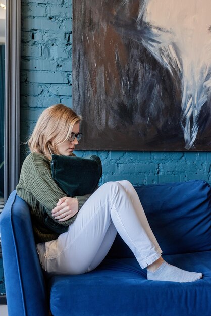 Jovem mulher pensativa caucasiana em suéter verde e calça jeans branca, sentada no sofá com os olhos fechados em uma elegante sala de estar azul