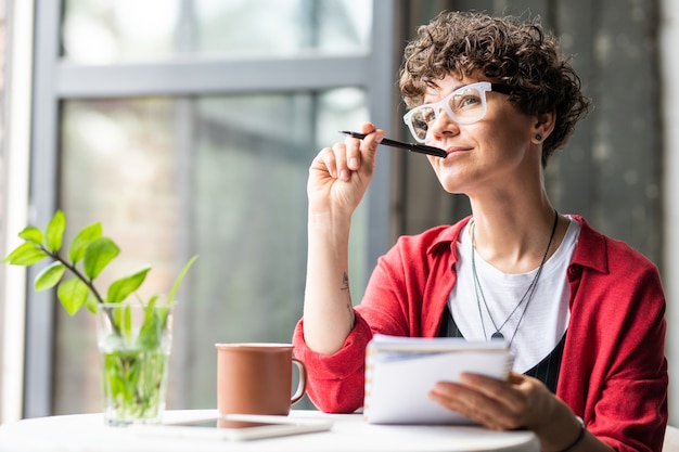 Foto jovem mulher pensativa em óculos pensando em novas ideias criativas enquanto está sentada à mesa no café ou no escritório