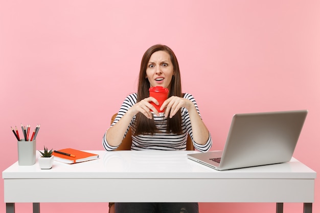 Jovem mulher perplexa, perplexa, segurando uma xícara de café ou chá, trabalhando em um projeto, sentada no escritório com o laptop do pc