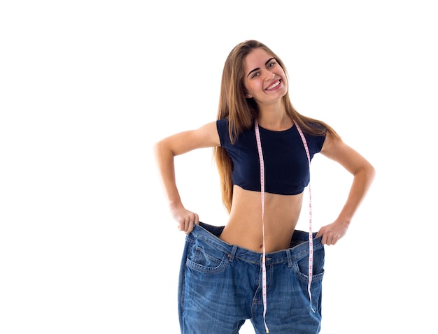 Foto jovem mulher positiva com centímetro em volta do pescoço segurando seu jeans de tamanho muito maior em fundo branco no estúdio