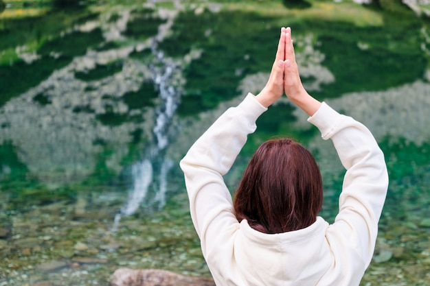 Jovem mulher praticando ioga e meditando na margem do lago com água transparente e cachoeira refletida nela. Férias e feriados nas montanhas. Relaxamento na natureza.