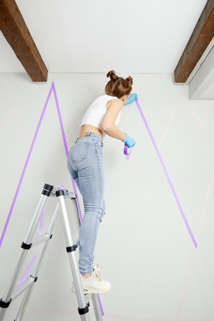 Jovem mulher preparando a parede para pintar em pé na escada, aplicando fita adesiva na parede do seu projeto