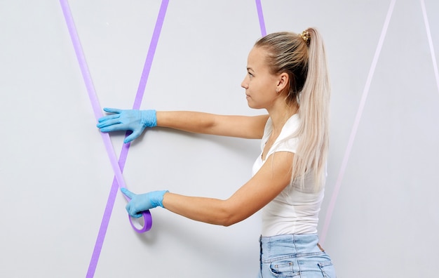 Jovem mulher preparando a parede para pintar em pé na escada, aplicando fita adesiva no projeto de diy da parede