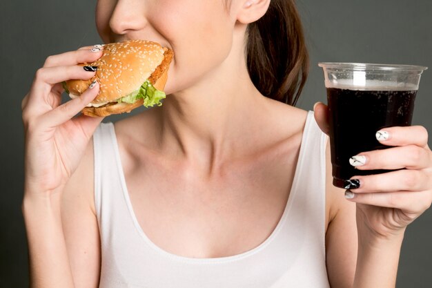 Jovem mulher que come o Hamburger no fundo cinzento.