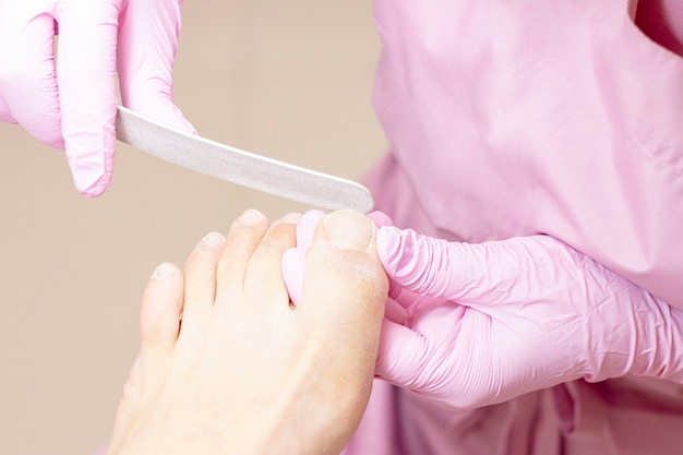 Jovem mulher que obtém o pedicure profissional em um salão de beleza, close up. as mãos do pedicuro em luvas de borracha protetora são aplicadas com unhas nas unhas usando uma lixa de unha. pedicure, conceito de manicure