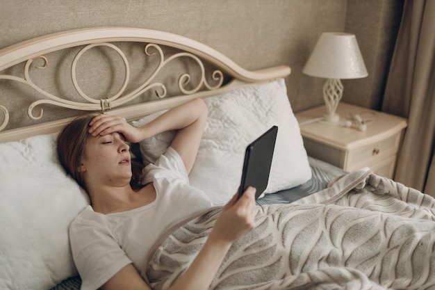 Jovem mulher que sofre com dor de cabeça, gripe, doença, frio, em casa, deitada na cama com tablet digital