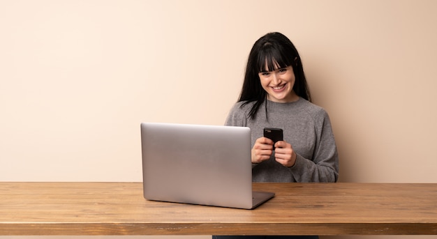 Jovem mulher que trabalha com seu laptop, enviando uma mensagem com o celular