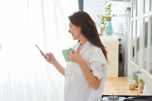 Jovem mulher que usa o smartphone que inclina-se na mesa de cozinha com caneca e organizador de café em uma casa moderna.