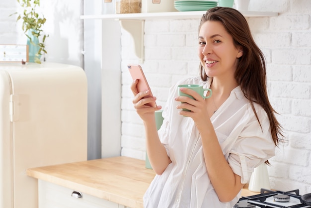 Jovem mulher que usa o smartphone que inclina-se na mesa de cozinha com caneca e organizador de café em uma casa moderna.