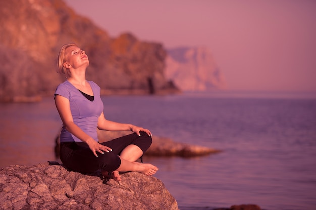 Jovem mulher relaxada caucasiana se senta em uma pedra e aprecia os raios do sol de verão no fundo do mar durante as tão esperadas férias em um dia de verão. Conceito de descanso e recuperação