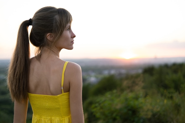 Jovem mulher relaxada em pé no campo verde, olhando a vista do pôr do sol na natureza à noite. Conceito de relaxamento e meditação.