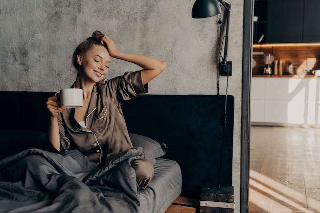 Jovem mulher relaxada em um terno elegante de casa acordando com uma xícara de café na cama em um quarto aconchegante e elegante em casa, estendendo-se da noite de sono e aproveitando o tempo de lazer matinal e o sol da janela