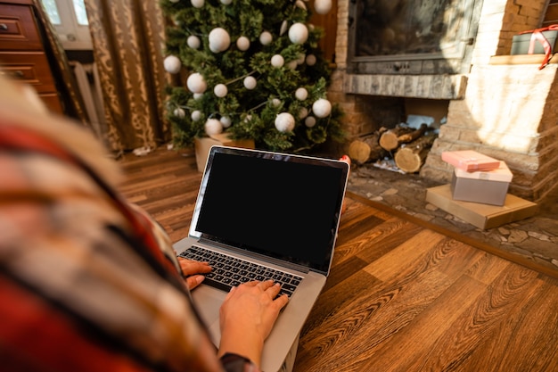 Jovem mulher relaxando em casa durante o Natal, sentada no chão da sala de estar em frente à árvore de Natal, navegando na internet em seu laptop.