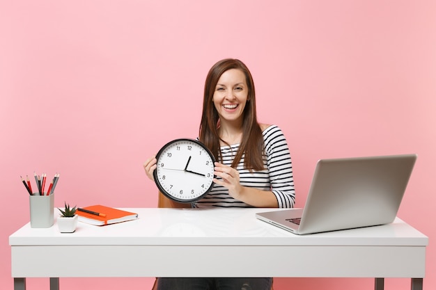 Jovem mulher rindo em roupas casuais segurando um despertador redondo, sentada no trabalho na mesa branca com um laptop pc contemporâneo