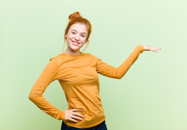 Jovem mulher ruiva bonita sorrindo, sentindo-se confiante, bem-sucedida e feliz, mostrando o conceito ou a ideia no espaço da cópia do lado contra a parede verde