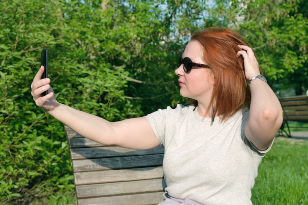 Jovem mulher ruiva com óculos de sol ajeita o cabelo para tirar uma foto de si mesma ao telefone. Mulher faz uma selfie em um smartphone em um parque da cidade.