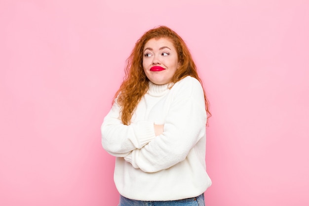 Jovem mulher ruiva encolher os ombros, sentindo-se confusa e incerta, duvidando com os braços cruzados e olhar intrigado na parede rosa